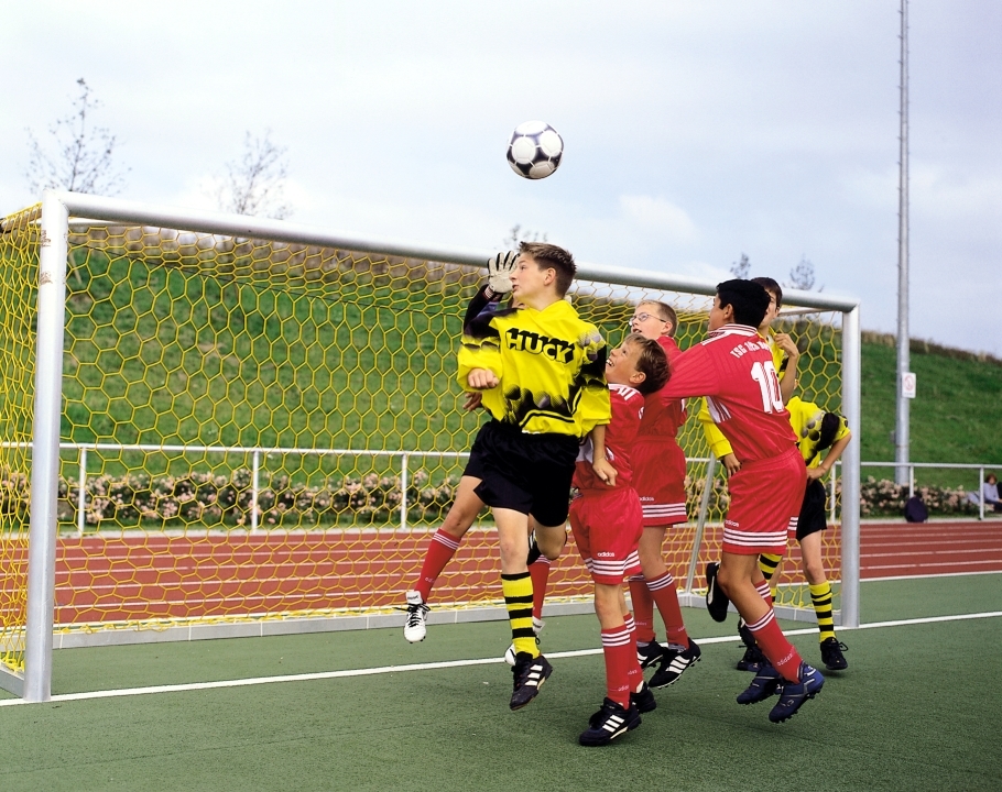 Mehrere jugendliche Fußballspieler kämpfen um den Ball bei einem Kopfballduell vor dem Tor. Ein Torwart in gelbem Trikot beobachtet die Situation.